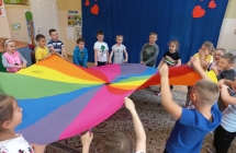 Uśmiechnięte dzieci z gr. VII podczas zabawy z kolorową chustą w sali.