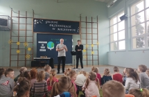 Dwoje mężczyzn, jeden z instrumentem muzycznym, drugi z mikrofonem i słuchająca publiczność - dzieci 