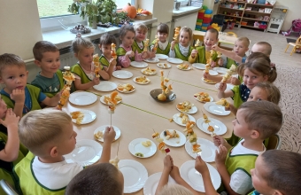 Czterolatki w zielonych fartuszkach siedza wokol  stolu i prezzentuja wykonane samodzielnie szaszlyki owocowe.