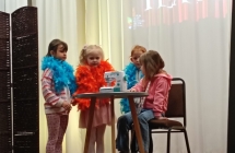 5. Trzy dziewczynki stoja w kolorowym boa, jedna siedzi przy stoliku, na ktorym stoi maszyna do szycia. Inscenizacja wiersza Kaczki
