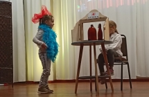 4. Jedna dziewczynka stoi w kolorowym szalu boa z duza kokarda na glowi, druka w bialym kitlu wyglada przez okienko z napisem apteka. Inscenizacja wiersza Kaczka Dziwaczka
