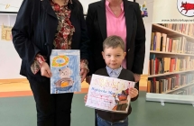 Chlopiec pozuje do zdjecia z dyplomem oraz swoja wyrozniona praca. Chlopiec stoi obok Pan organizatorek konkursu.