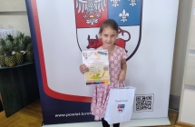 Dziewczynka z dyplomem i nagrodami na tle napisu.
